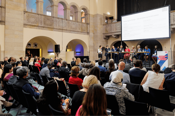 Menschen in einem großen Saal verfolgen eine Podiumsdiskussion über Partnerschaften für Veränderung.