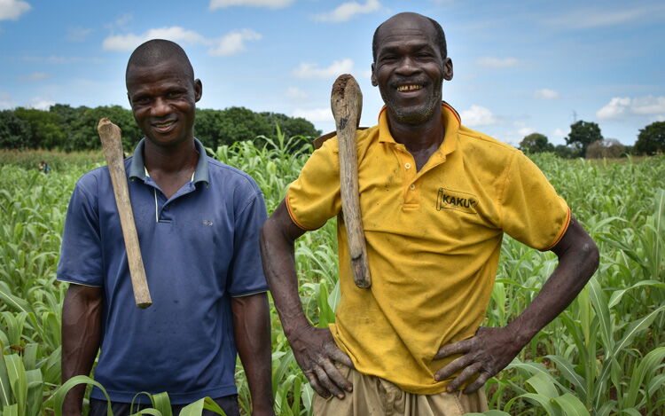 Zwei Bauern stehen lächelnd mit Werkzeugen auf ihren Schultern in einem grünen Feld unter blauem Himmel.