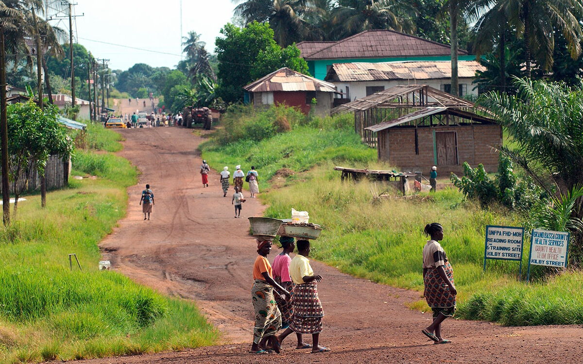 People walk along a red dirt road in a rural village, with greenery and buildings lining the path under a clear sky.