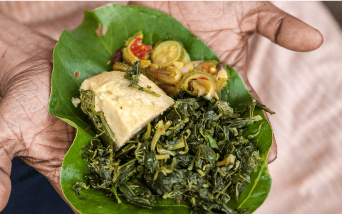 Eine Hand hält ein Blatt mit gekochtem Gemüse, einem Stück Tofu und Gewürzen, die für ein traditionelles Gericht arrangiert sind.