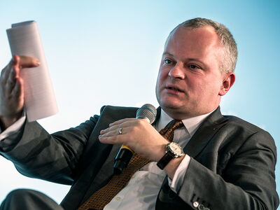 Ein Mann im Anzug spricht in ein Mikrofon und hält ein Dokument, während er in einem Interview oder einer Diskussion ist.