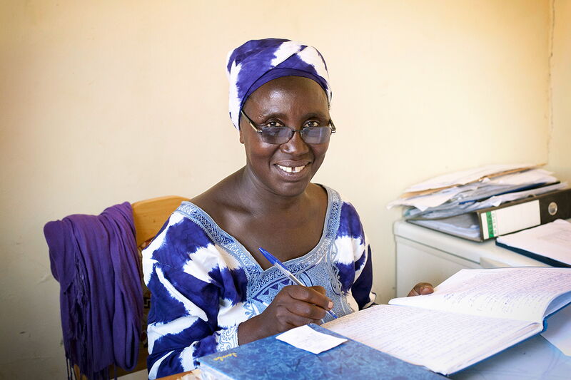 A smiling woman in traditional blue and white attire writes in a notebook at a desk, embodying a sense of determination.