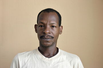 A man in a light shirt looks directly at the camera against a plain beige background.