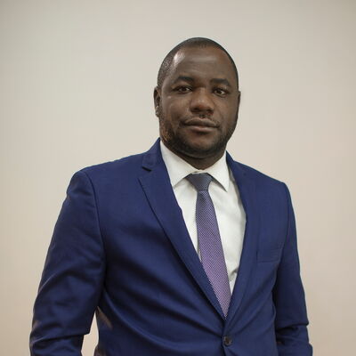 Ein Mann in einem blauen Anzug steht selbstbewusst vor einem neutralen Hintergrund mit den Händen in den Taschen.