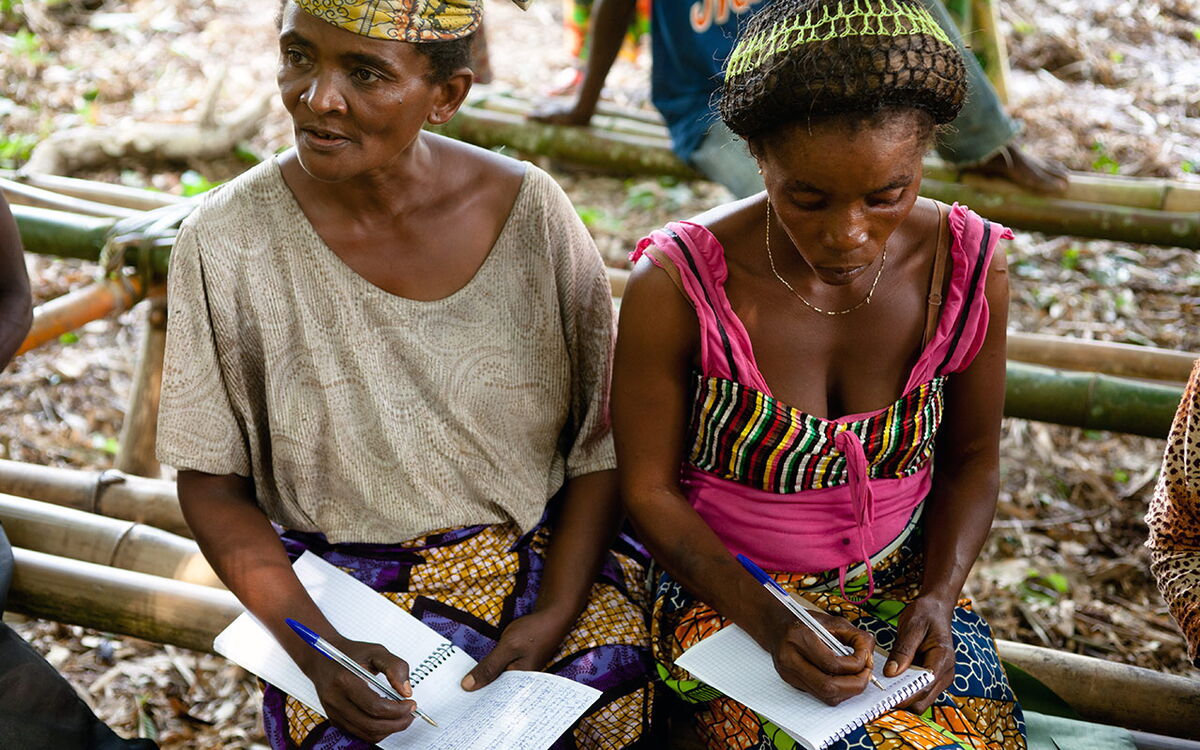 Zwei Frauen sitzen auf Bambusbänken im Freien und machen sich Notizen in Hefte während eines Workshops.