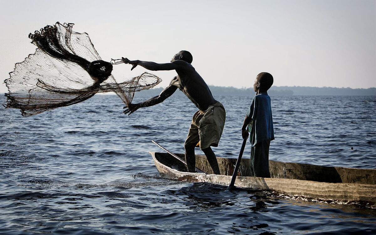A man skillfully casts a fishing net from a wooden boat as a child watches on a serene lake.