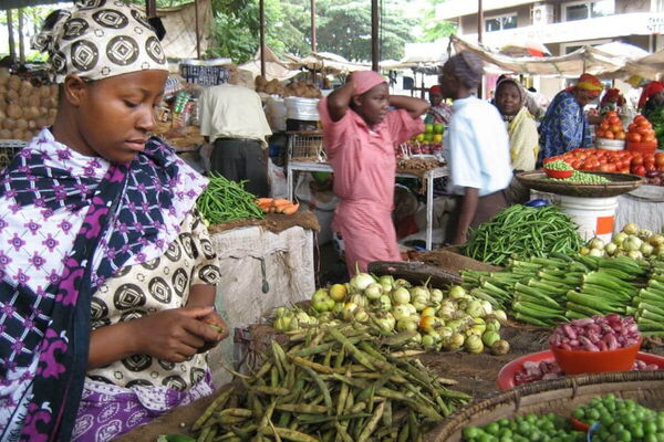Eine Frau sortiert Gemüse auf einem belebten Marktstand, umgeben von bunten frischen Produkten und anderen Verkäufern.