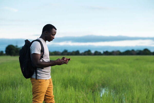 Ein Mann steht in einem grünen Feld und benutzt sein Smartphone, während er einen schwarzen Rucksack trägt.