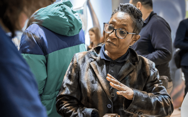 Eine Frau in einem gemusterten Mantel spricht gestikulierend mit einer anderen Person in einer belebten Umgebung.