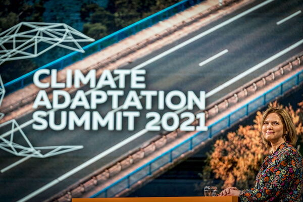 Eine Frau steht vor einem großen Bildschirm mit der Aufschrift "Climate Adaptation Summit 2021" und lächelt in die Kamera.