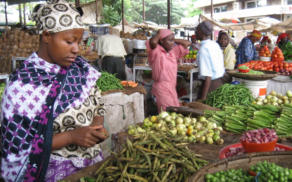 Eine Frau in traditioneller Kleidung verkauft frisches Gemüse auf einem geschäftigen Markt in Tansania.