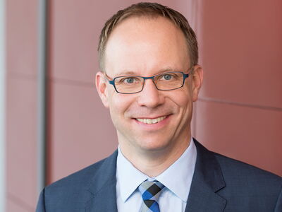 Ein Mann in einem blauen Anzug lächelt vor einer roten Wand in die Kamera.