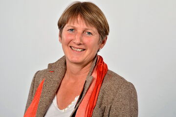 A woman in a brown jacket and orange scarf smiles warmly against a plain background.