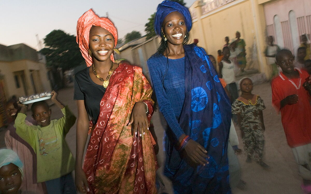 Zwei Frauen in traditionellen Kleidern lächeln, während sie mit Kindern durch eine lebhafte Straße spazieren.