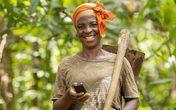 A smiling farmer in Togo holds a mobile phone, standing in lush greenery with a hoe resting on her shoulder.