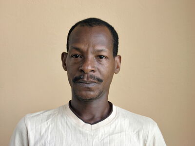 Ein Mann in einem cremefarbenen Hemd schaut ernst in die Kamera vor einem beigen Hintergrund.