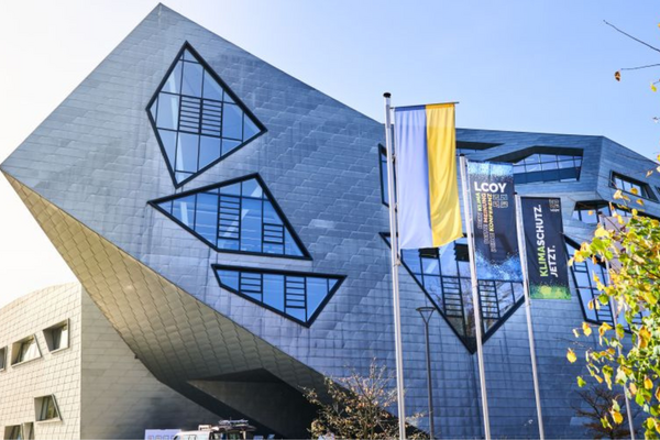 Ein futuristisches Gebäude mit geometrischen Fenstern und LCOY-Banner, flankiert von Fahnen, steht im Herbstlicht.