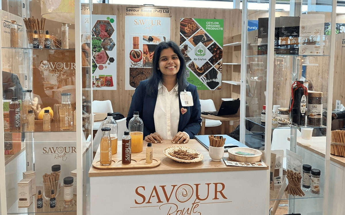 Eine Frau steht lächelnd an einem Messestand von Savour Route mit Getränken und Gewürzen.