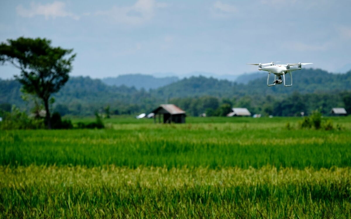 Eine Drohne fliegt über ein grünes Feld vor einer hügeligen Landschaft mit verstreuten Hütten.