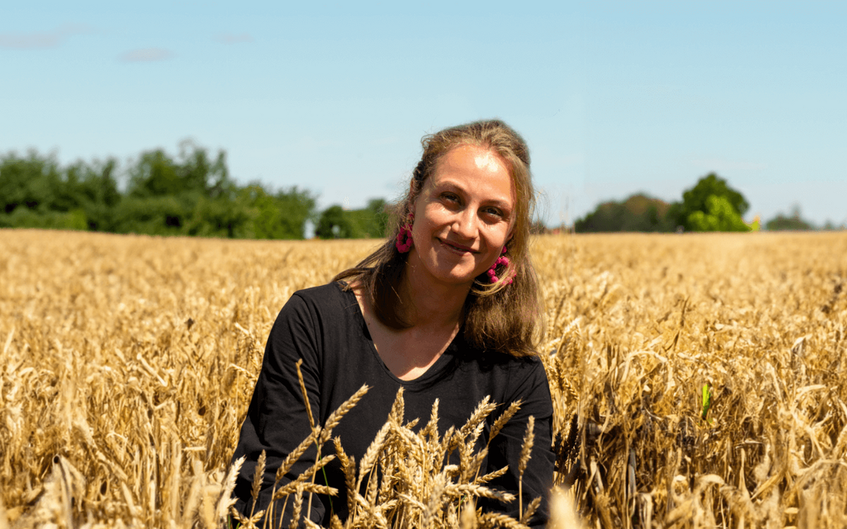 Eine Frau mit rosa Ohrringen lächelt in einem goldenen Weizenfeld unter blauem Himmel.