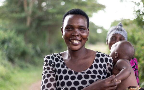 Eine lächelnde Frau in gepunkteter Bluse hält ein Baby im Arm, während eine weitere Person im Hintergrund steht.
