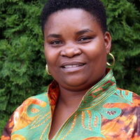 Eine Frau mit buntem Oberteil und goldenen Ohrringen lächelt vor einem Hintergrund aus grünen Sträuchern.