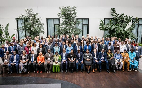 Eine große Gruppe von Menschen posiert gemeinsam für ein Gruppenfoto vor einem Gebäude mit Pflanzen im Hintergrund.