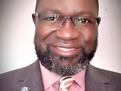 Ein lächelnder Mann in Anzug und Krawatte trägt eine Brille und hat einen gepflegten Bart.