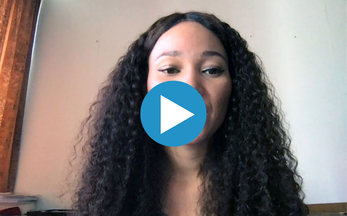Eine Frau mit langen, lockigen Haaren spricht in die Kamera, während ein Play-Button das Video kennzeichnet.
