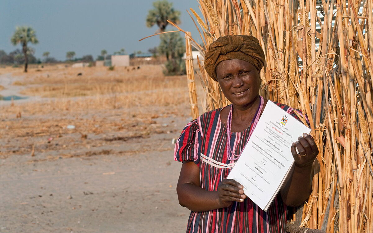 Eine Frau in traditioneller Kleidung hält eine Landrechtsurkunde in Namibia vor einem Feld in der Hand.