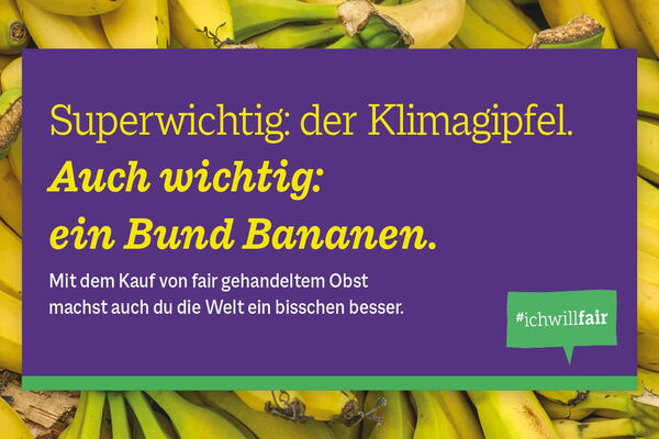Ein Bündel Bananen liegt hinter einem Schild mit dem Text zur Bedeutung fair gehandelter Produkte und dem Hashtag #ichwillfair.