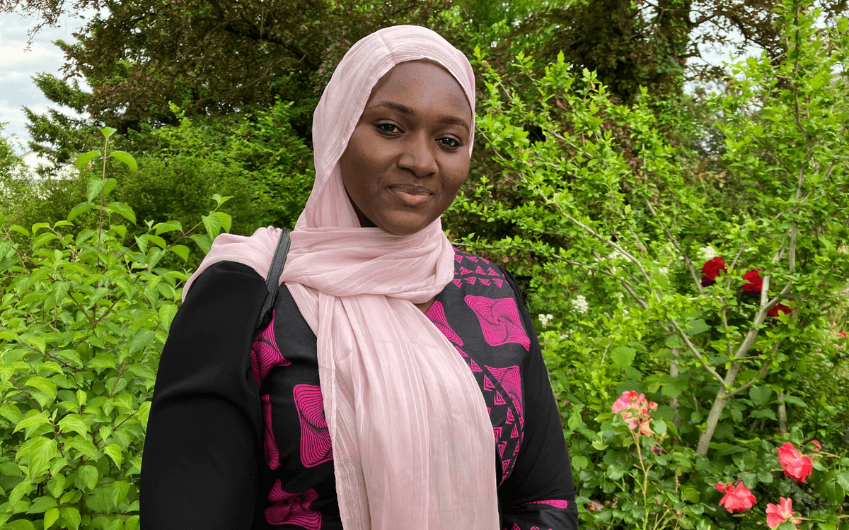 Eine Frau in einem rosa Kopftuch und schwarzem Kleid steht lächelnd vor üppigem Grün und blühenden Rosen.
