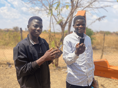 Two men smile while holding mobile phones outdoors on a sunny day, standing near a fence and a tree.