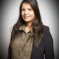 Eine Frau in einem dunklen Blazer steht vor einem neutralen Hintergrund und lächelt in die Kamera.