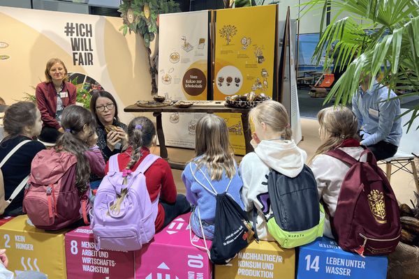 Kinder mit Rucksäcken sitzen vor einem Stand, der den Prozess von Kakao zu Schokolade erklärt, während eine Frau spricht.