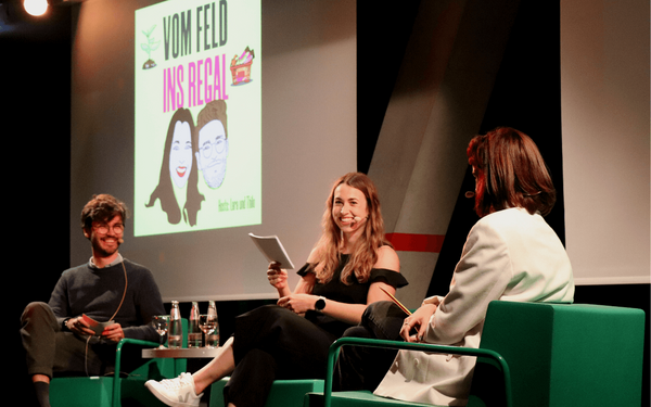 Drei Personen diskutieren auf einer Bühne während eines Podcasts mit dem Titel "Vom Feld ins Regal".