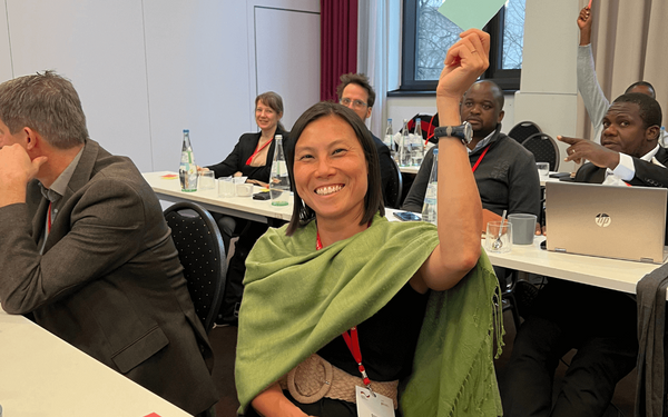 Eine lächelnde Frau hebt in einem Konferenzraum die Hand, während sie an einer Diskussion teilnimmt.