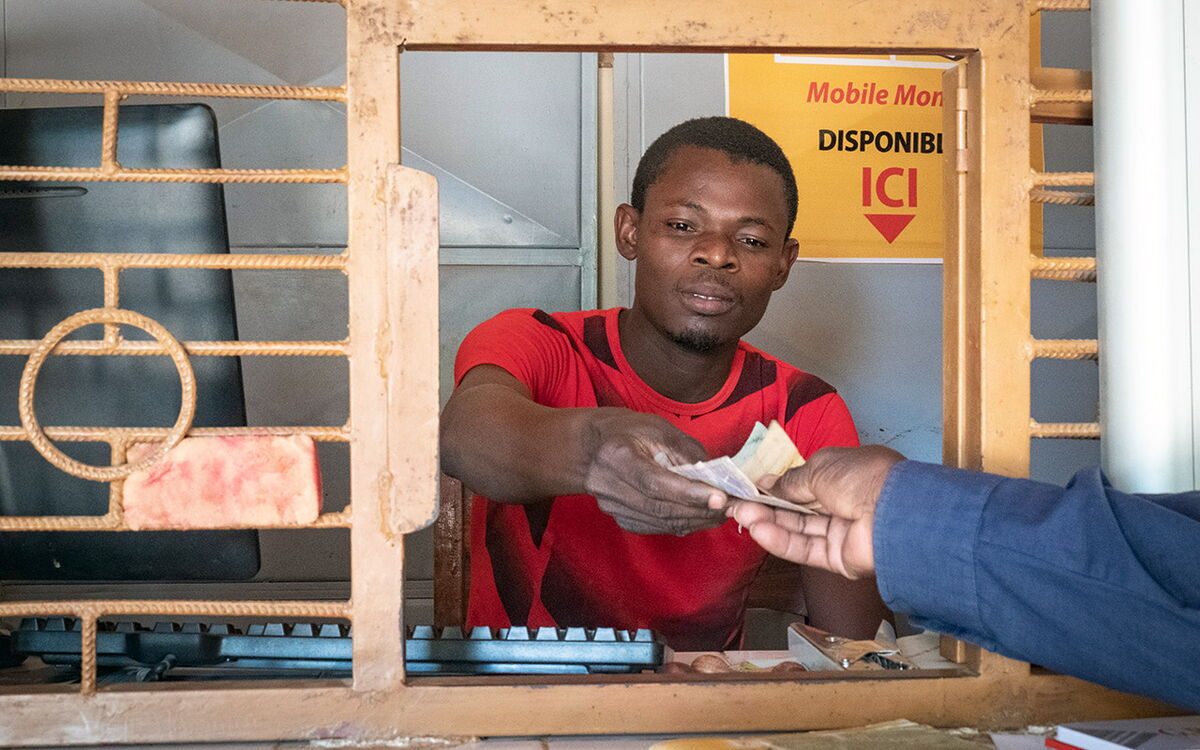 Ein Mann am Schalter in rotem Hemd überreicht durch ein Gitter hindurch Geld an eine andere Person.