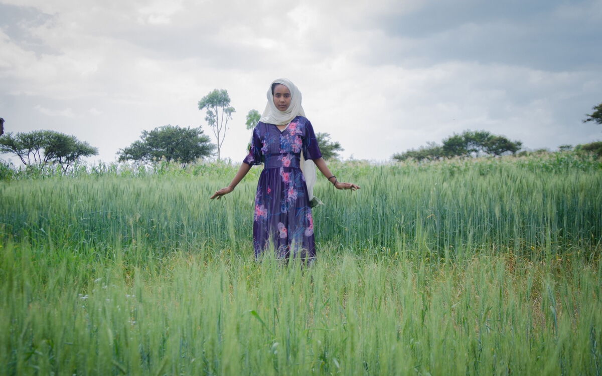 Eine Frau in einem blumigen Kleid steht in einem grünen Feld unter bewölktem Himmel und hebt leicht die Arme.