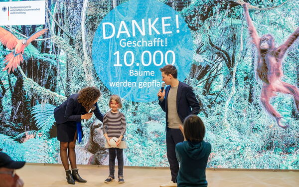 A woman and a man celebrate with a child in front of a backdrop announcing the success of planting 10,000 trees.
