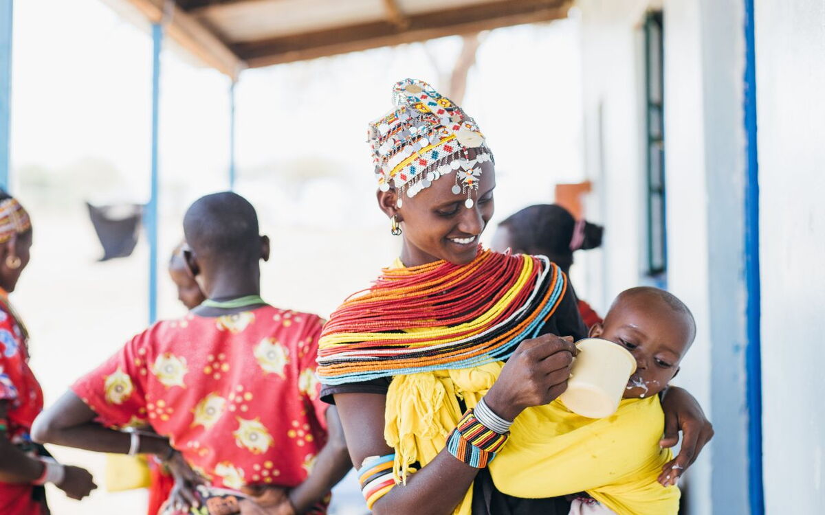 Eine Frau in traditioneller Kleidung lächelt, während sie einem Kind aus einer Tasse zu trinken gibt.