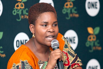 A person in a vibrant patterned outfit speaks into a microphone at a "do agric" event, highlighting agricultural initiatives.