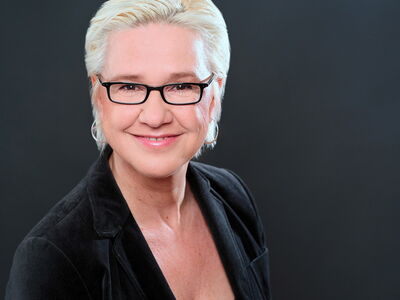 Eine lächelnde Frau mit Brille und hellem Haar trägt einen schwarzen Blazer und blickt freundlich in die Kamera.