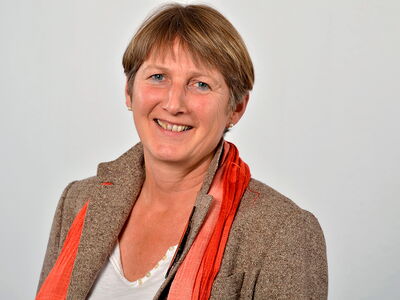 Eine lächelnde Frau in einem braunen Blazer und rotem Schal blickt freundlich in die Kamera.