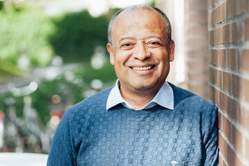 A smiling man in a blue sweater stands against a brick wall, radiating warmth and approachability.