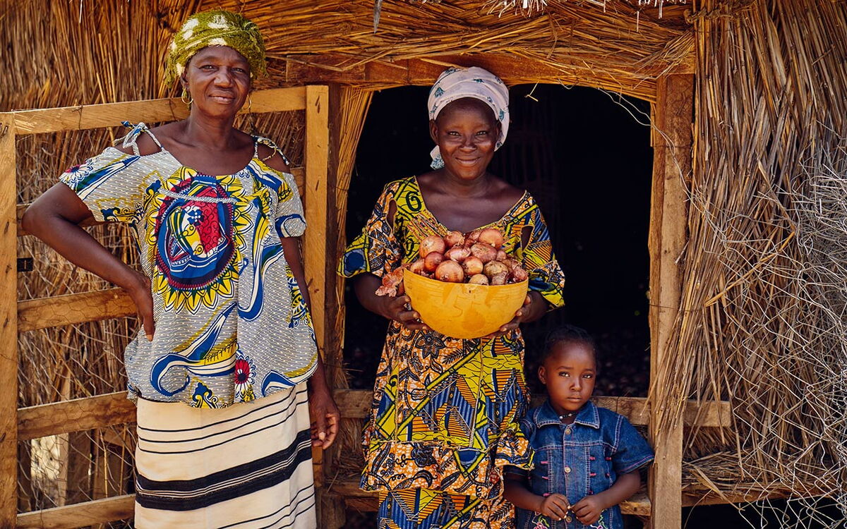 Zwei Frauen in bunter Kleidung stehen mit einem Kind vor einer Hütte, eine hält eine Schüssel voller Zwiebeln.