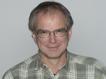 A man with glasses smiles warmly, wearing a plaid shirt against a neutral background.