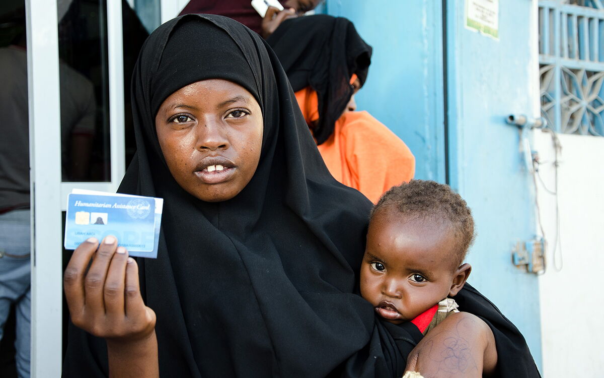 Eine Frau in schwarzem Hijab hält einen Gutschein und trägt ein Kind vor einer blauen Tür.