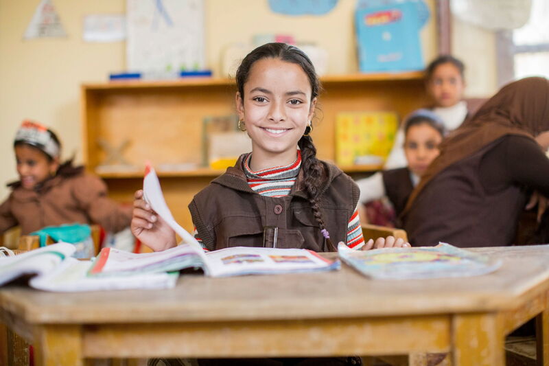 A smiling girl in a classroom sits at a wooden desk, surrounded by open books and classmates in the background.