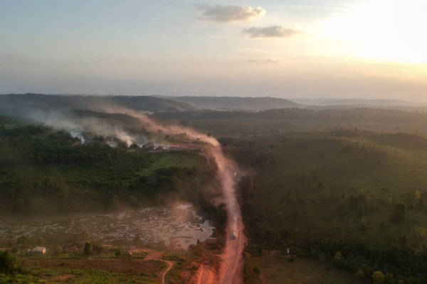 Ein staubiger Weg schlängelt sich durch eine grüne Landschaft, während Rauchfahnen in der Abenddämmerung aufsteigen.